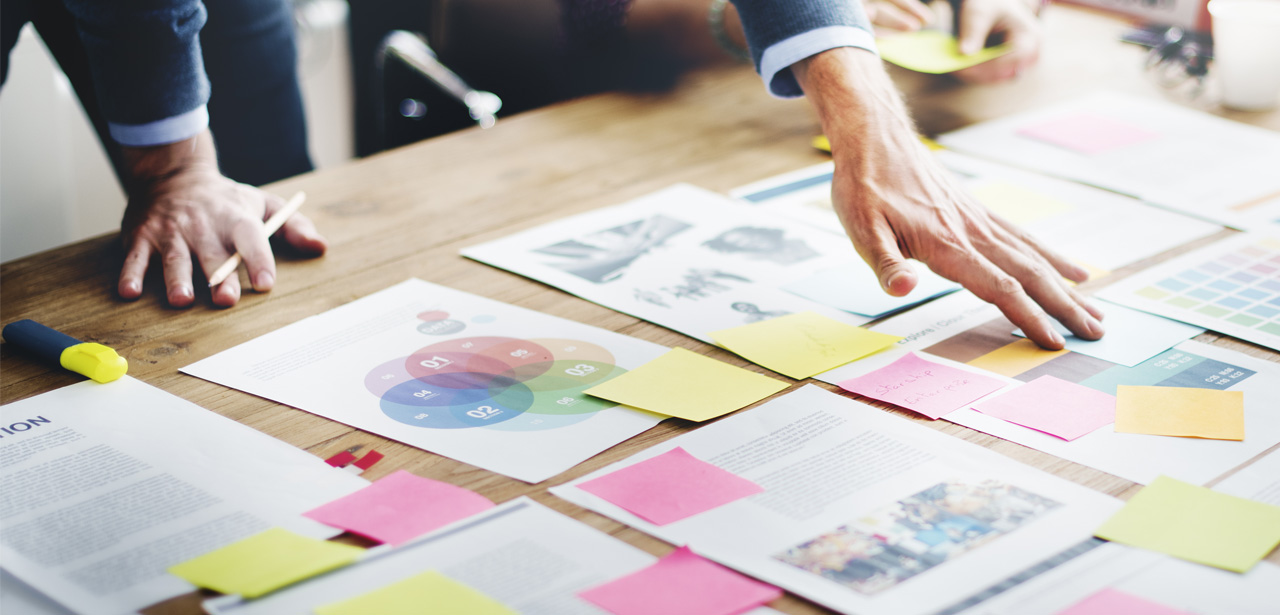 Visuel plusieurs feuilles et post-it posés sur une table en bois avec 2 personnes les organisant